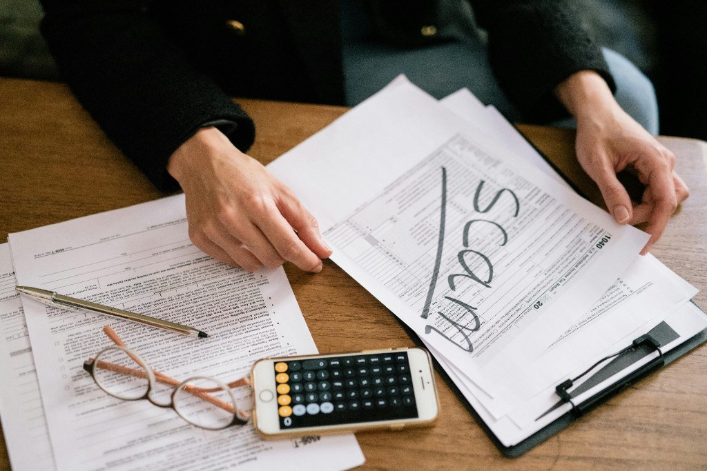 Hands holding a tax document marked SCAM on a desk with paperwork, calculator phone, and glasses, illustrating employment and tax-related fraud